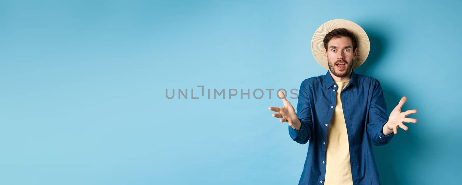 Surprised young man in summer hat, raising hands up staring with disbelief and amazement at camera, impressed with big event, standing on blue background.