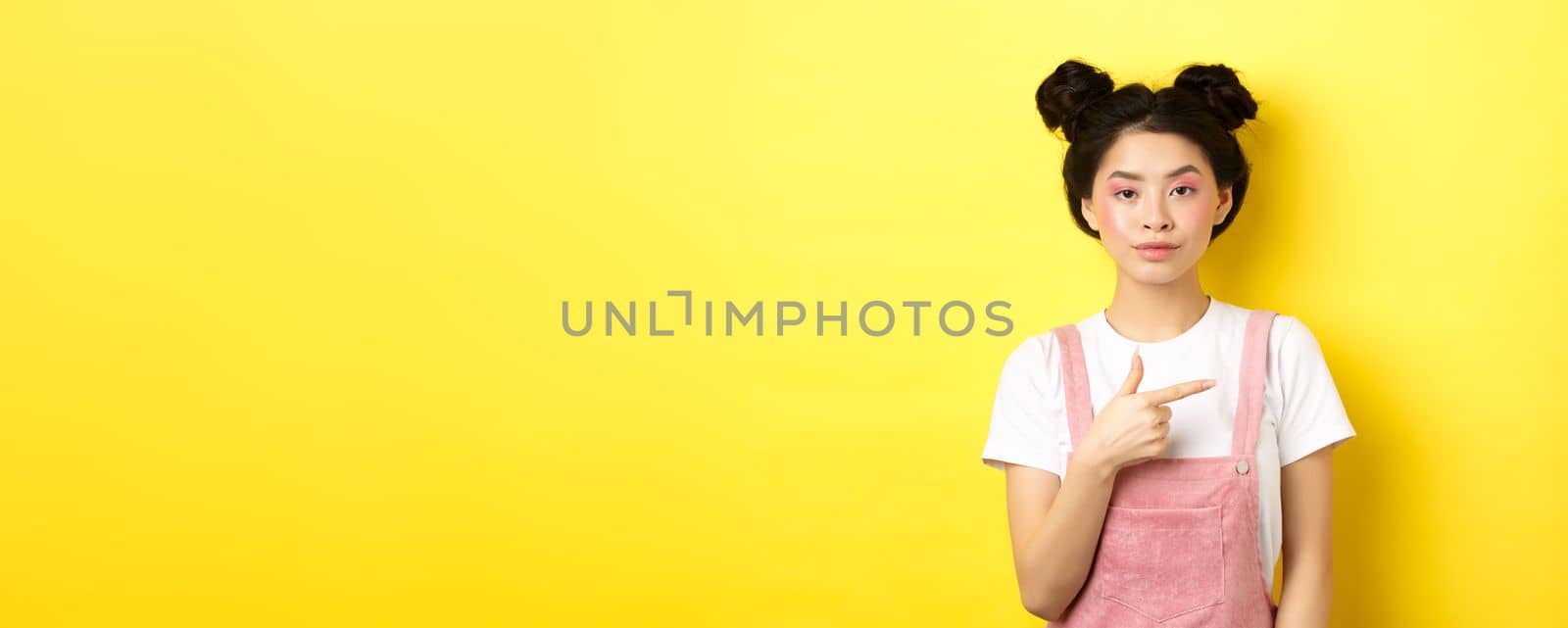 Stylish asian teen girl with makeup and summer clothes, pointing finger right and look serious, standing against yellow background.