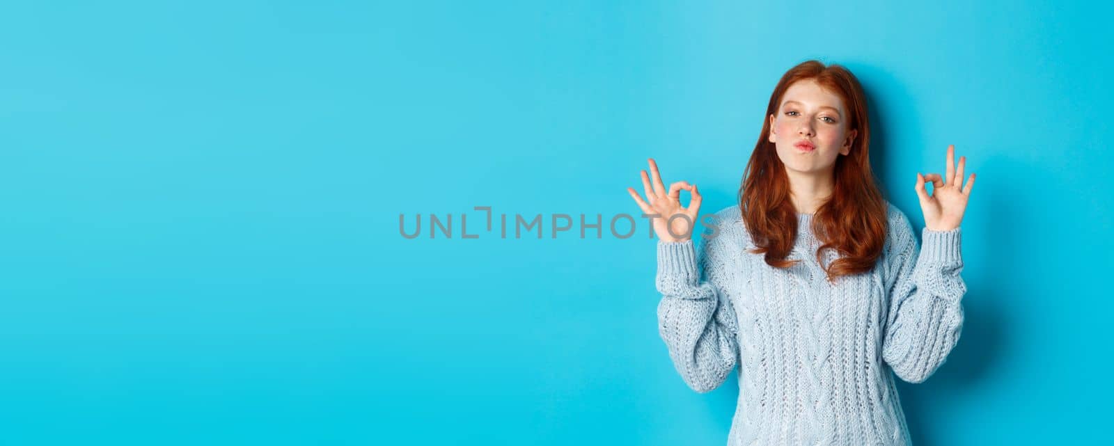 Redhead teenage girl showing okay signs, looking satisfied and proud, agree, give positive answer, praising good choice, standing over blue background.