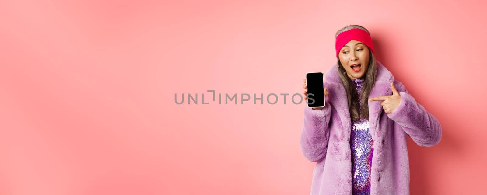 Online shopping and fashion concept. Cool asian granny in stylish faux fur coat pointing finger left at blank smartphone screen, checking out internet promo, pink background.