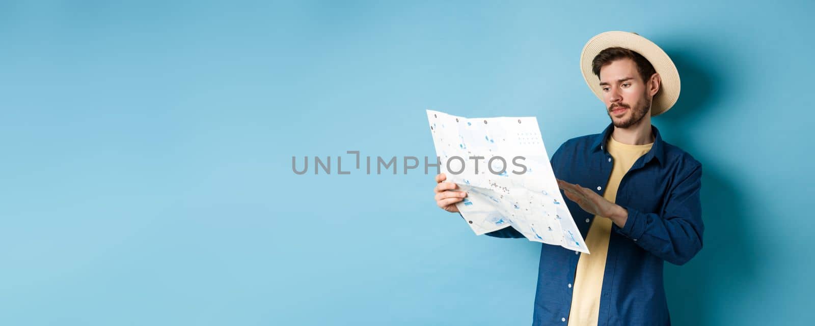Image of handsome caucasian man looking at travel map, studying route during summer vacation, standing in straw hat on blue background by Benzoix