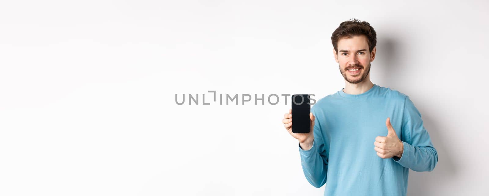 Smiling young man showing empty smartphone screen and thumbs up, praising good application, recommend mobile app, standing over white background by Benzoix