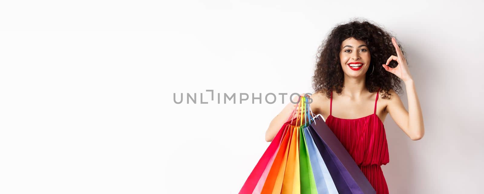 Beautiful woman in elegant dress, holding shopping bags and showing okay sign, recommend store with discounts, white background.