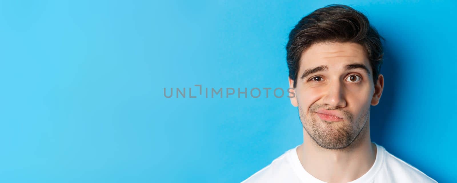 Headshot of skeptical guy looking at something unamusing, grimacing and standing reluctant against blue background.