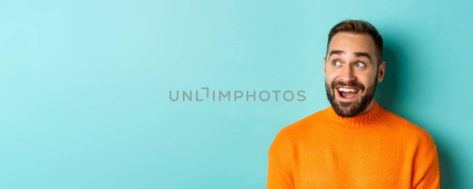 Close-up of handsome caucasian man smiling, looking left with surprised face, staring at logo, wearing orange sweater, standing against turquoise background.