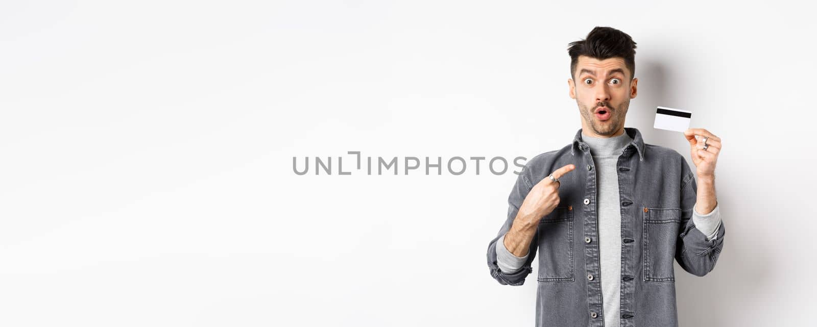 Wow look here. Excited guy pointing at plastic credit card and look amazed with awesome deal, standing against white background.
