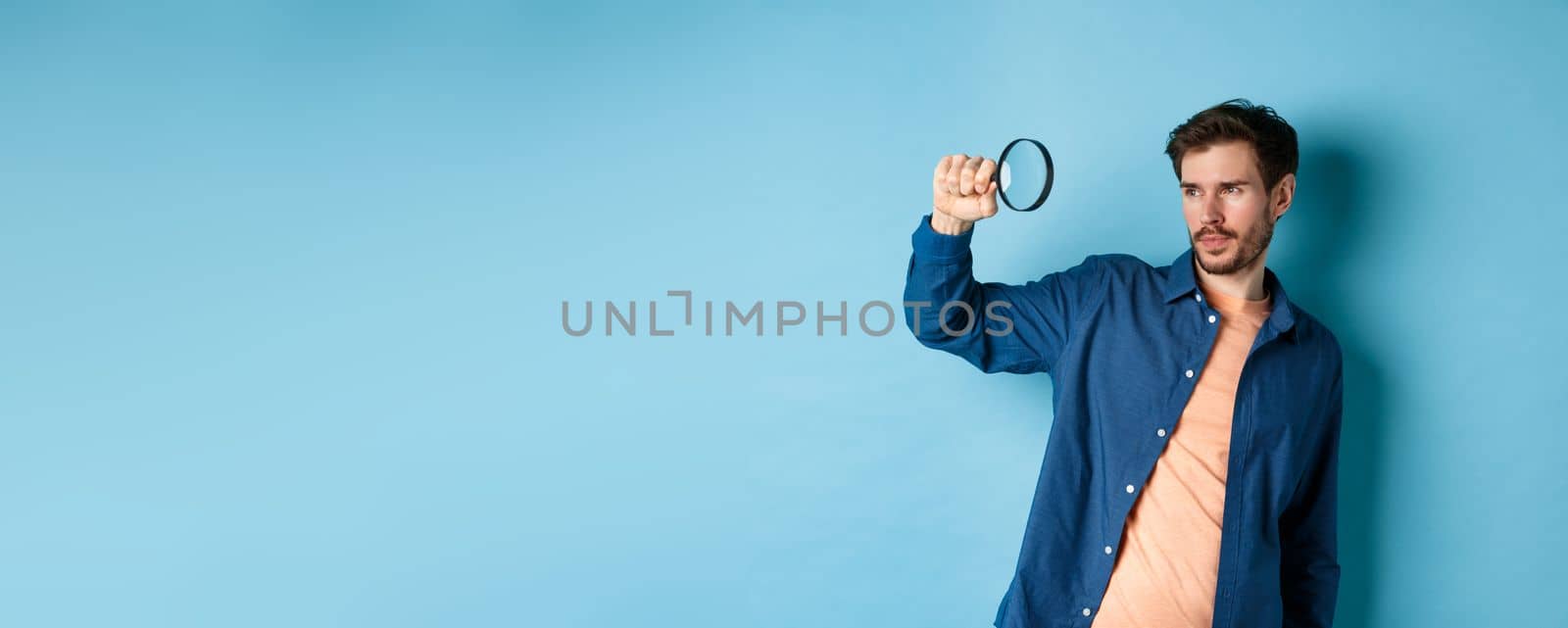 Image of young man searching for something, looking at empty space with magnifying glass, standing on blue background.