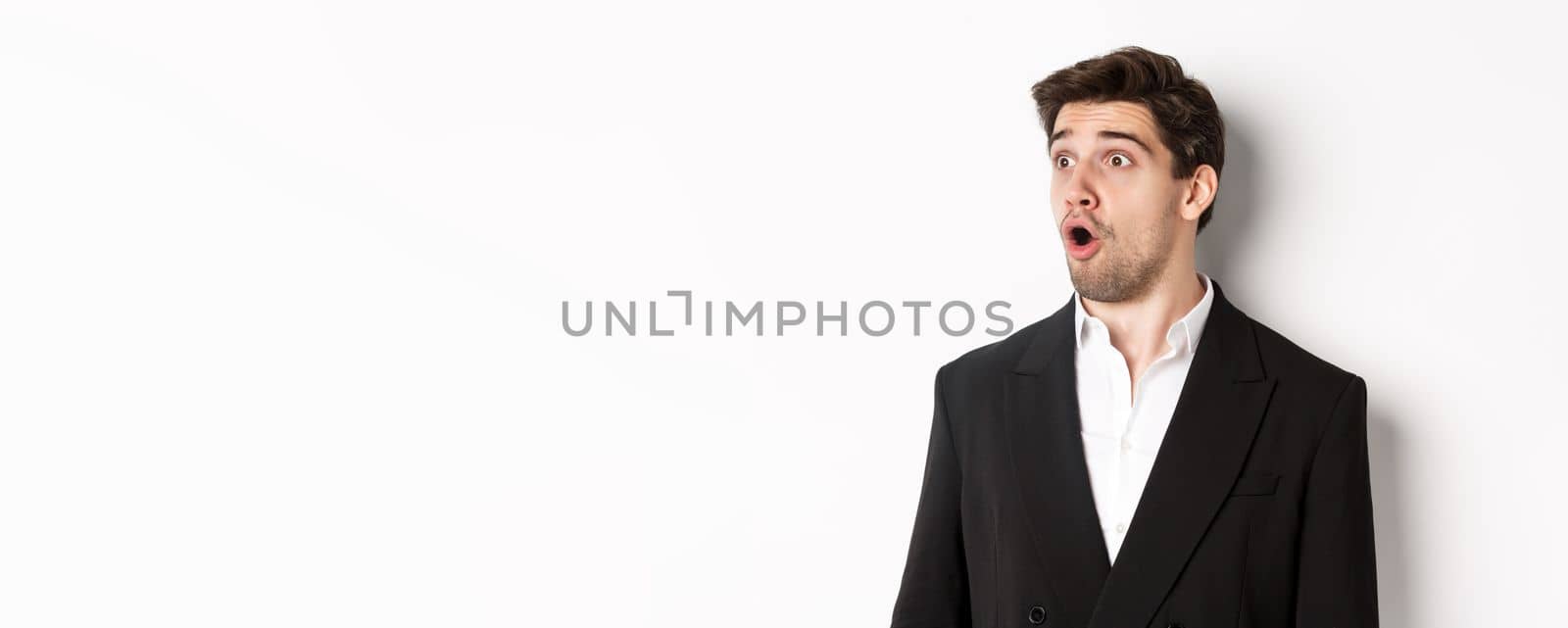 Close-up of impressed guy in trendy suit, open mouth and looking left wondered, standing against white background by Benzoix