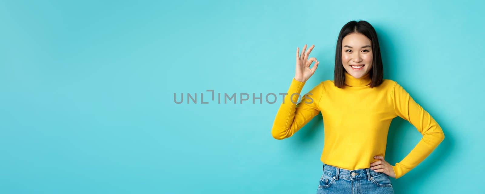 Happy beautiful asian woman smiling, showing okay gesture in approval, saying yes, standing pleased over blue background.