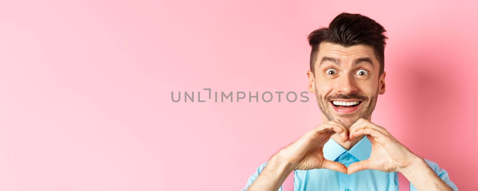 Valentines day concept. Close up of romantic guy looking happy, smiling and showing heart gesture, standing on pink background.