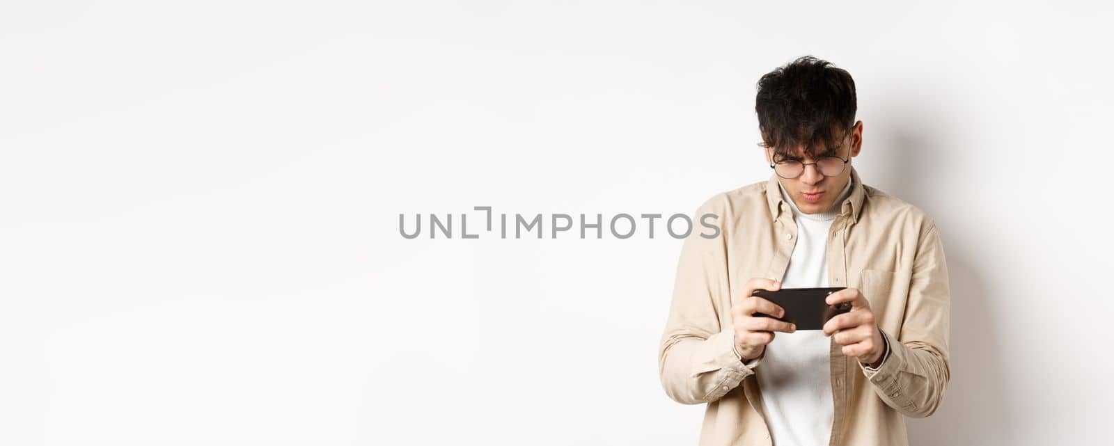 Handsome young man stare at mobile screen and playing video game on smartphone, standing on white background. Copy space
