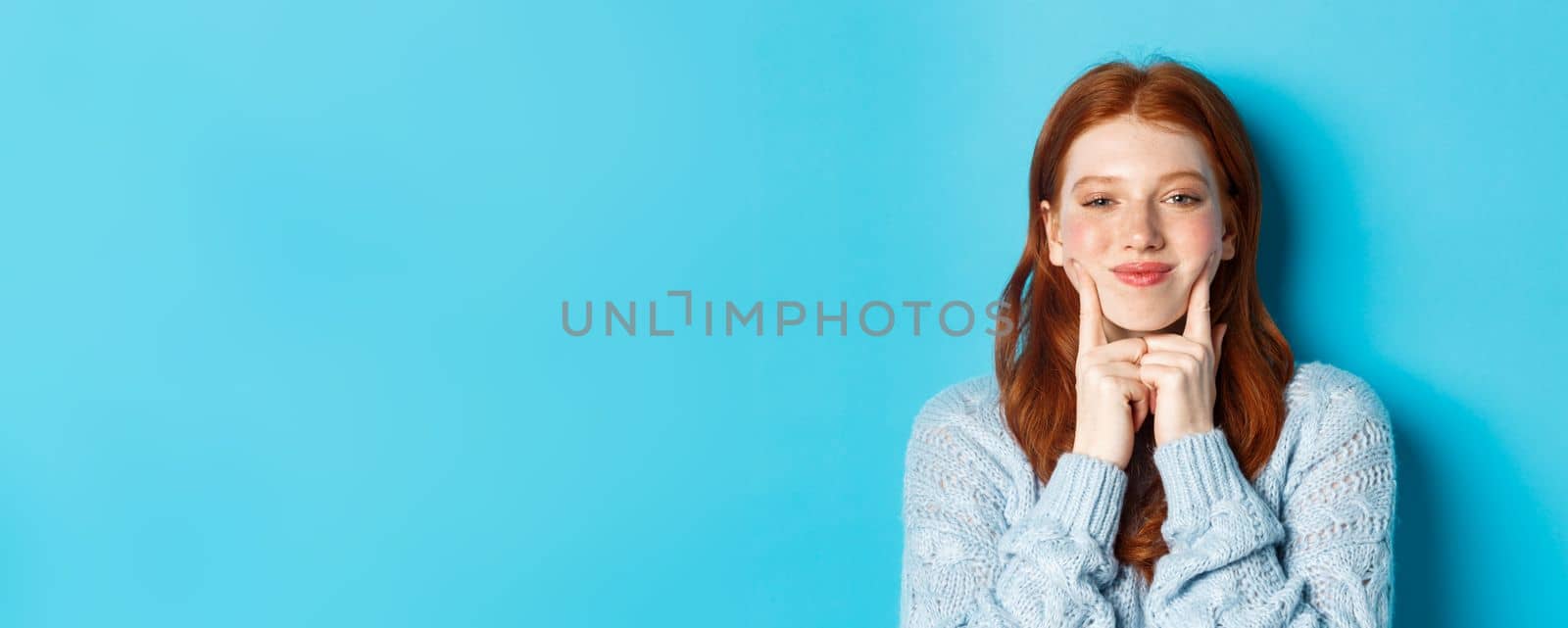 Close-up of cute and silly girl with red hair poking cheeks, showing dimples and smiling, standing against blue background by Benzoix