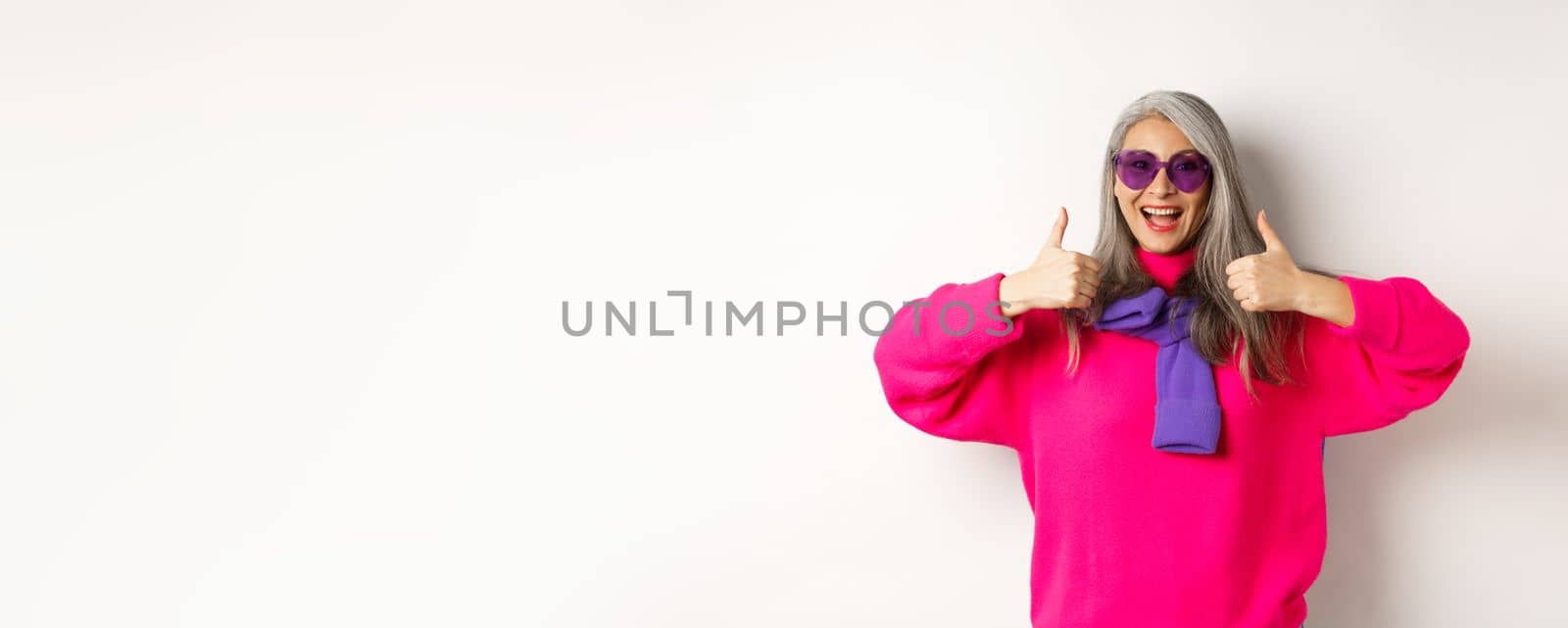 Shopping concept. Stylish asian senior woman in sunglasses and trendy outfit, showing thumb-up in approval, recommending shop, standing over white background by Benzoix