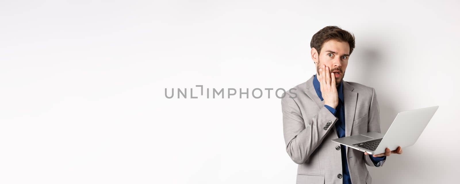 Shocked and troubled businessman have trouble on laptop, looking worried at camera, made mistake at work, standing on white background in suit.
