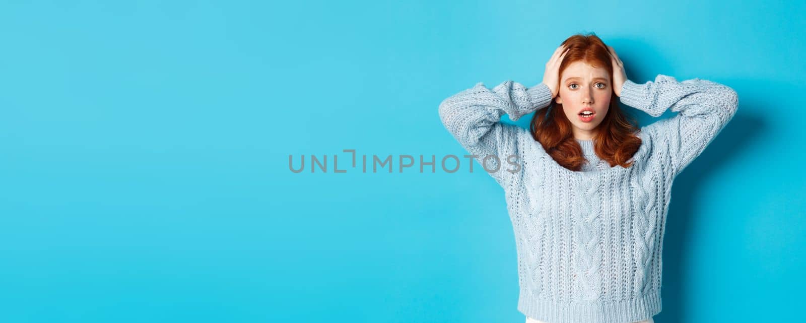 Nervous redhead girl standing overwhelmed, holding hands on head in panic and staring at camera, standing anxious against blue background by Benzoix