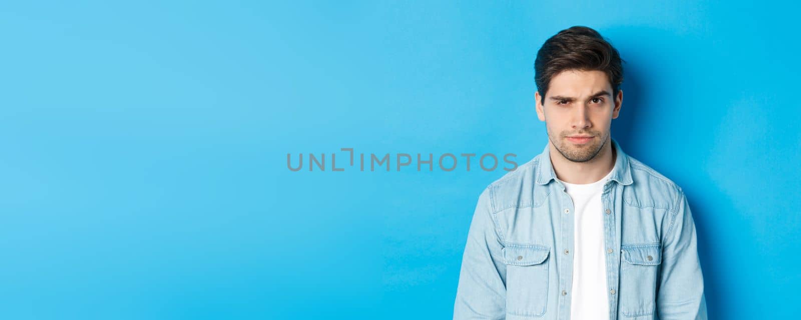 Close-up of suspicious and serious guy raising one eyebrow, looking at camera, standing against blue background.