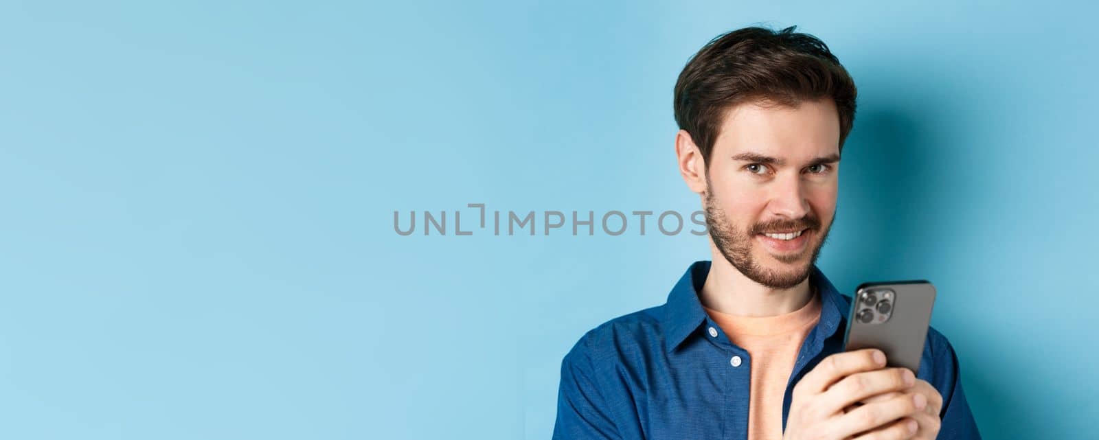 Close-up of attractive caucasian man holding mobile phone and smiling at camera, standing in casual outfit on blue background.