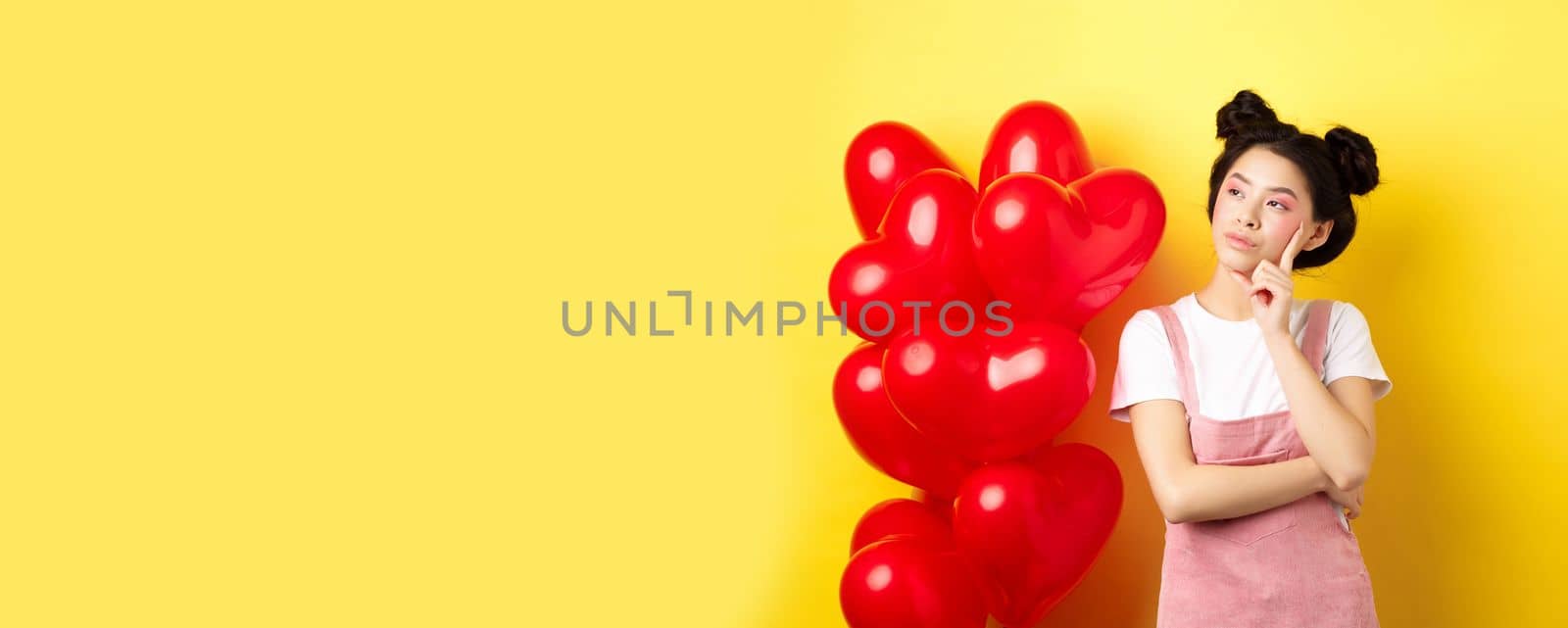 Valentines day concept. Pensive asian woman looking left, thinking about romantic date, standing near red hearts balloons on yellow background.