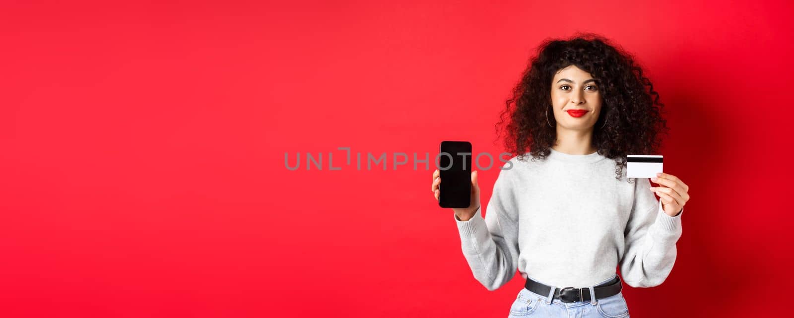 Young modern woman with curly hair showing plastic credit card and mobile phone screen, demonstrating online shopping app, standing on red background.