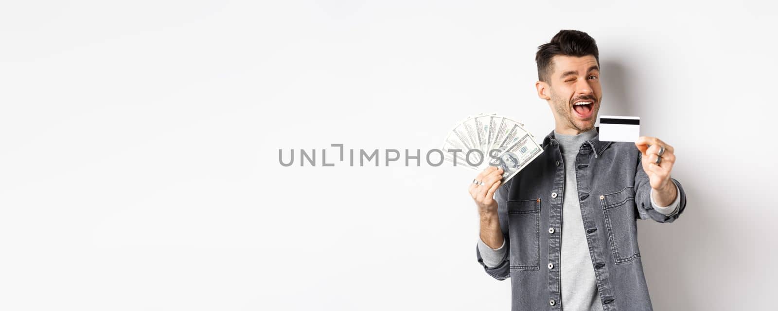 Image of handsome guy holding dollar bills but suggesting use plastic credit card, smiling friendly at camera, standing on white background by Benzoix