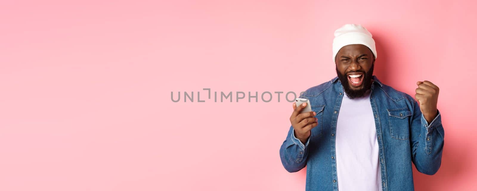 Technology and online shopping concept. Happy Black man rejoicing, winning in app, holding smartphone and scream yes, standing over pink background.