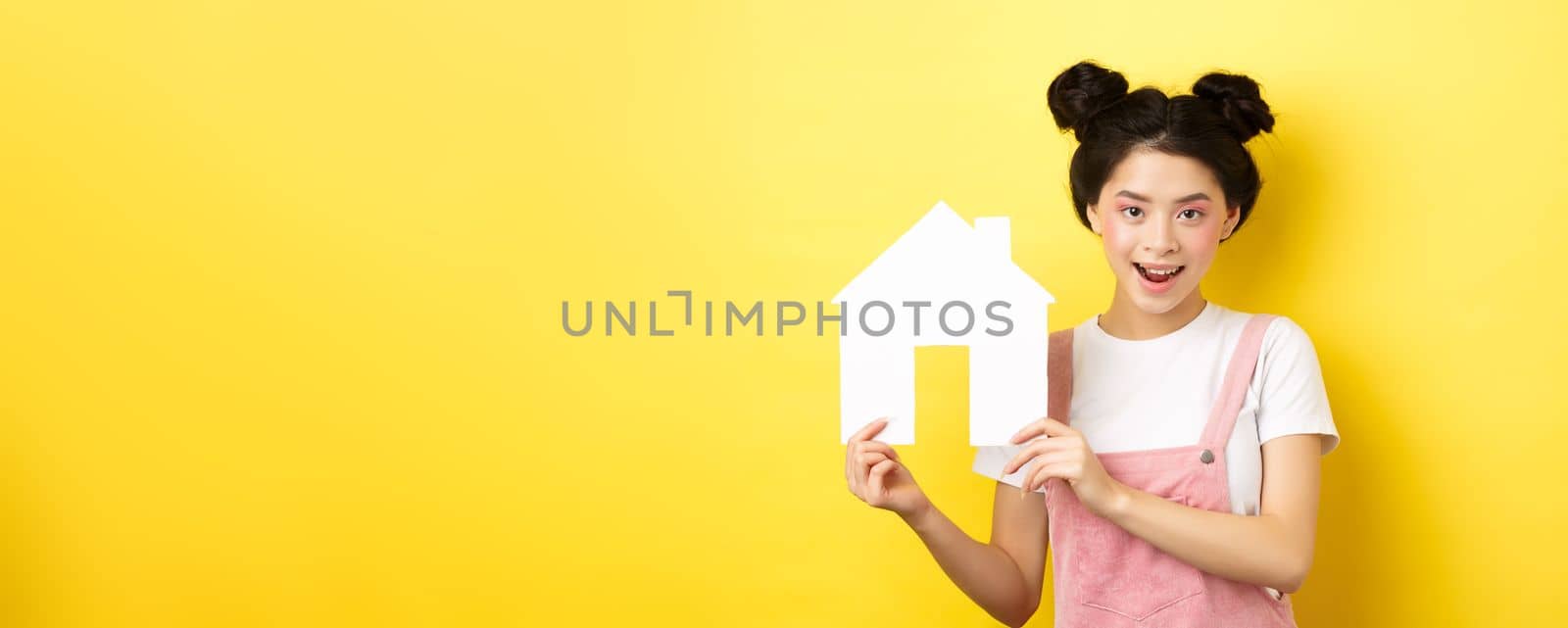 Real estate and family concept. Cute asian woman with bright makeup and stylish hairbuns, showing paper house cutout, smiling determined, yellow background by Benzoix