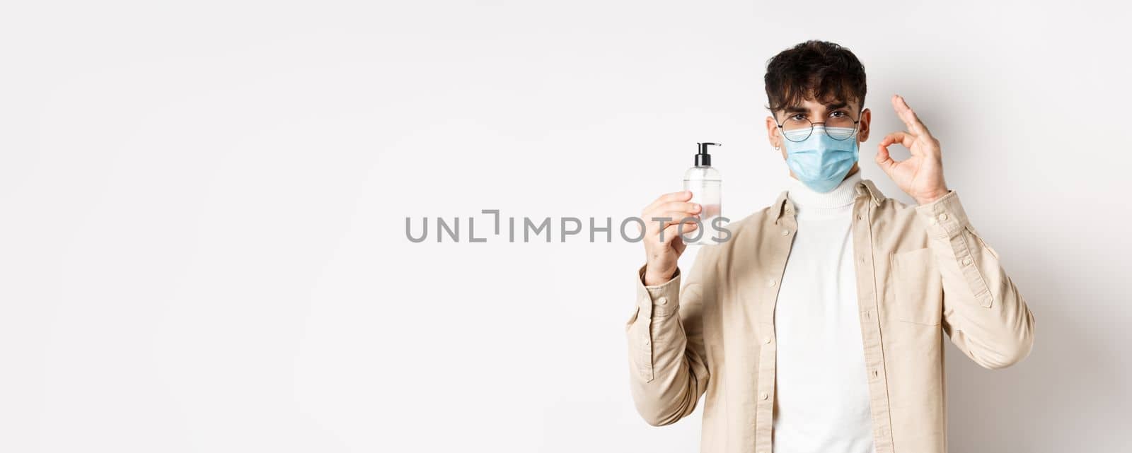 Health, covid and quarantine concept. Portrait of natural guy in glasses and face mask showing bottle of good hand sanitizer, make okay sign, recommending product, white background.