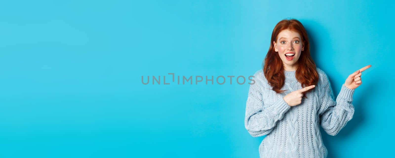 Winter holidays and people concept. Curious teenage girl in sweater, pointing fingers right and staring at camera amazed, showing promo offer, standing over blue background.