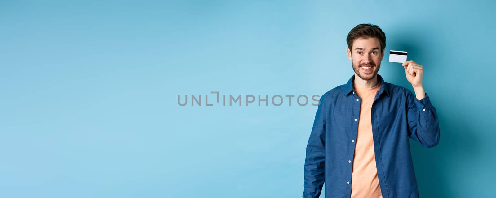 Happy caucasian guy smiling, showing plastic credit card, standing in casual clothes on blue background.