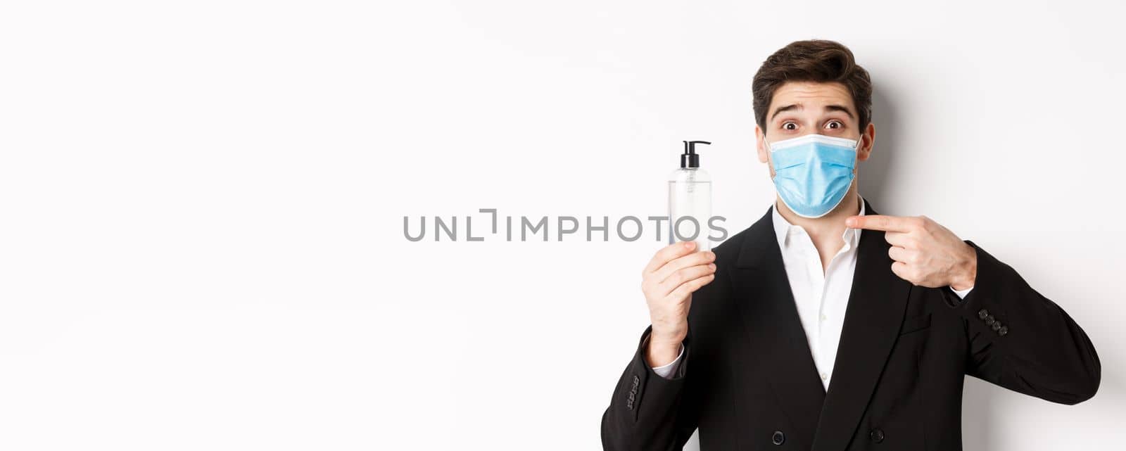 Concept of covid-19, business and social distancing. Close-up of handsome man in trendy suit and medical mask, recommending hand sanitizer, standing against white background.