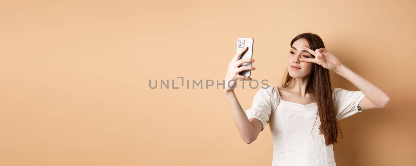 Cute woman taking selfie on smartphone, showing v-sign near eye, posing for social media photo, standing on beige background.