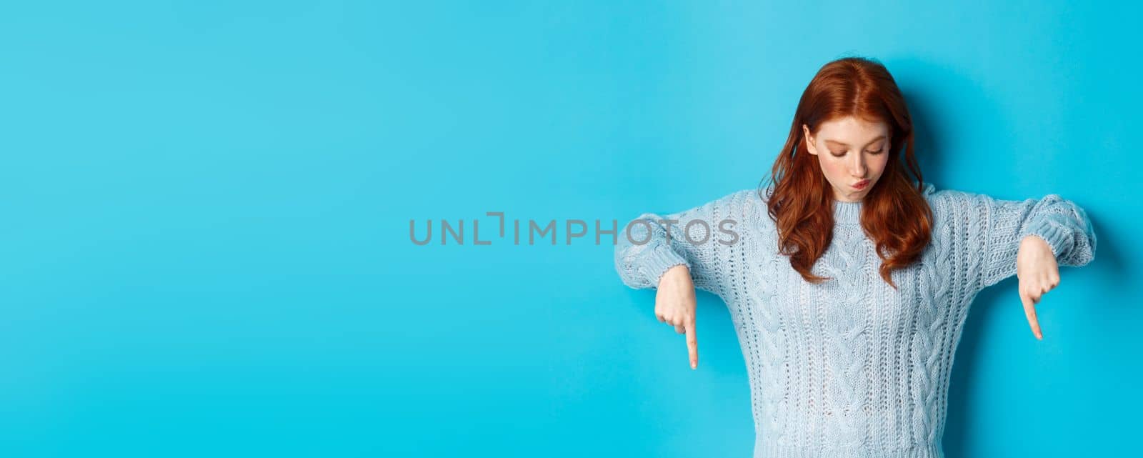 Winter holidays and people concept. Intrigued redhead girl, pointing and looking down thoughtful, making choice, standing over blue background.