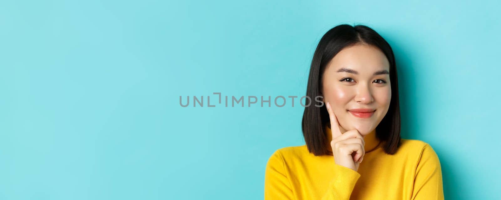 Beauty and makeup concept. Close up of thoughtful asian woman looking pleased at camera and smile, having an idea, standing over blue background by Benzoix