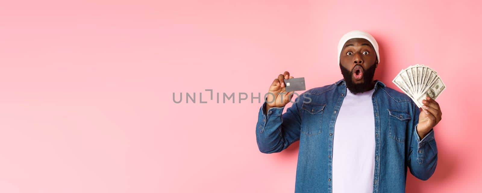 Shopping concept. Excited african-american man showing credit card and dollars, got deposit or money loan, standing over pink background by Benzoix