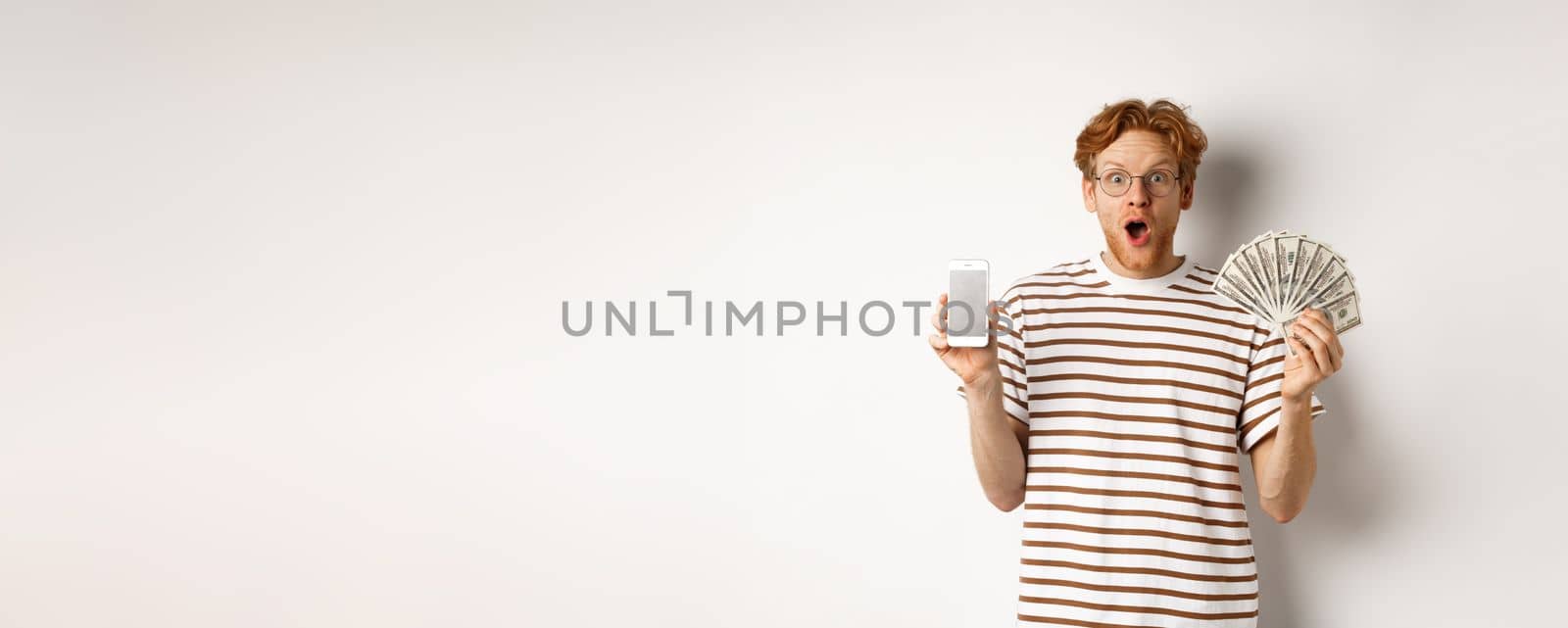 Amazed redhead man showing smartphone app on blank screen and money, winning prize cash online, standing over white background.