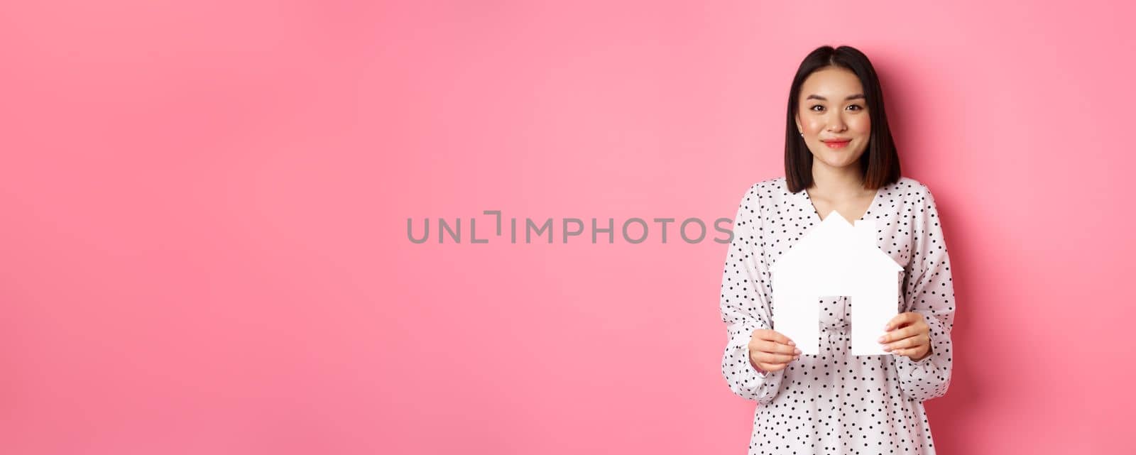 Real estate. Adult asian woman searching for home, holding house model and smiling, promo of broker company, standing over pink background.