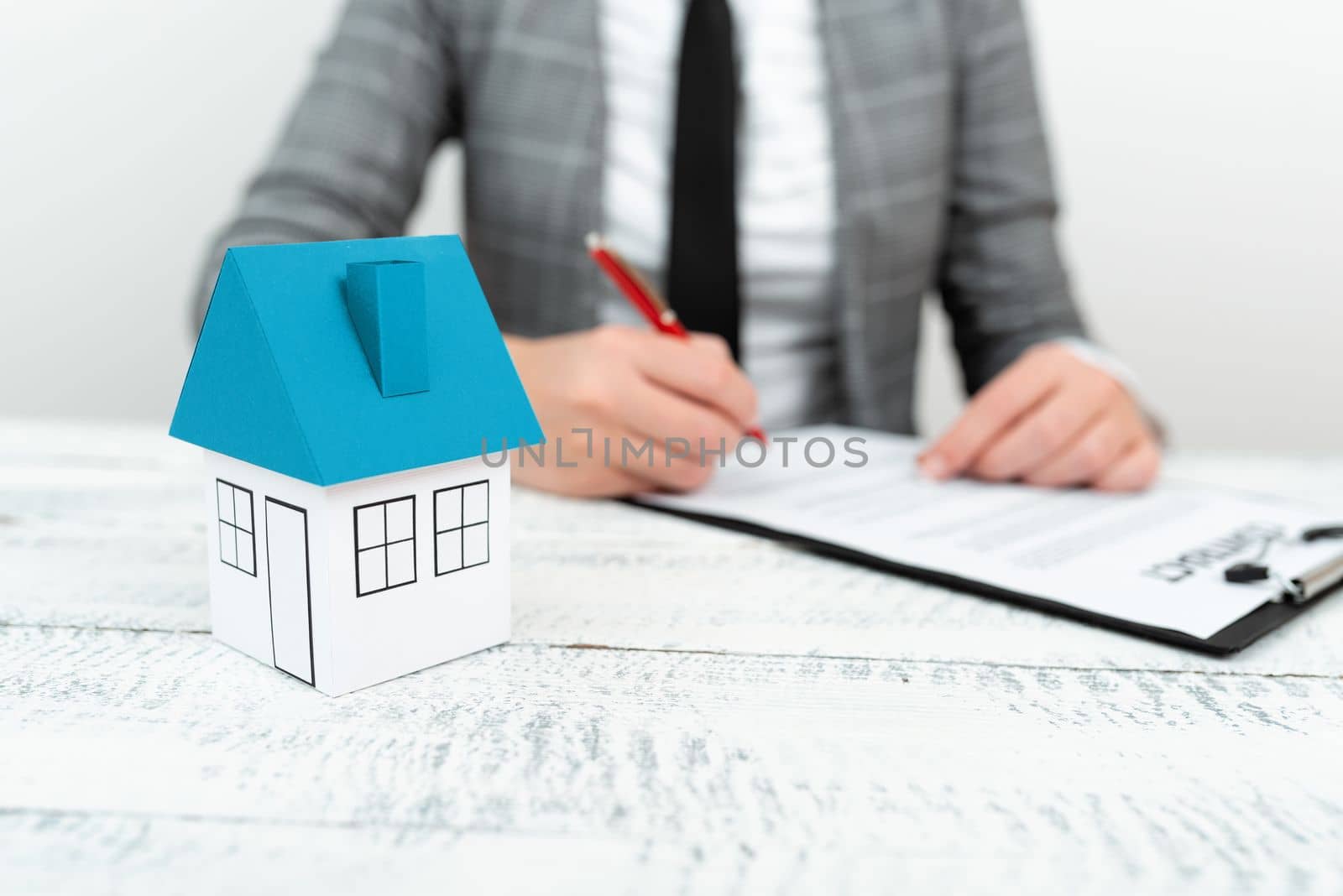 Businesswoman sitting and writing in notebook. Paper house on desk.