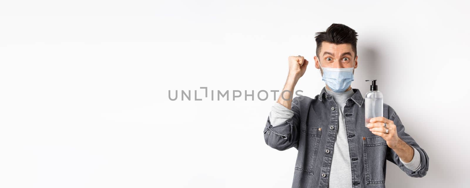 Health, covid and pandemic concept. Happy guy motivated to get rid from virus germs, showing bottle of hand sanitizer and fist pump, cheering while standing on white background.