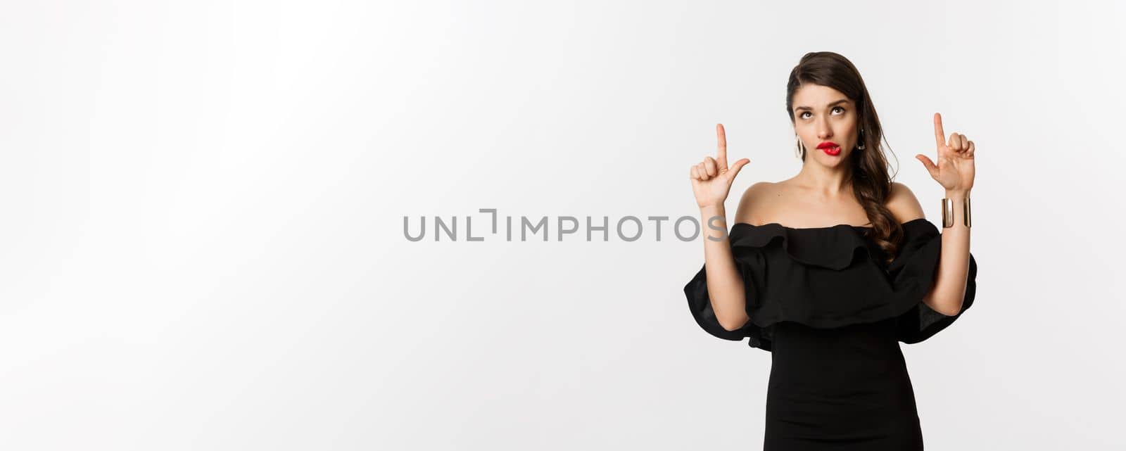 Fashion and beauty. Thoughtful attractive woman in black dress staring and pointing up, thinking with tempted look, white background.