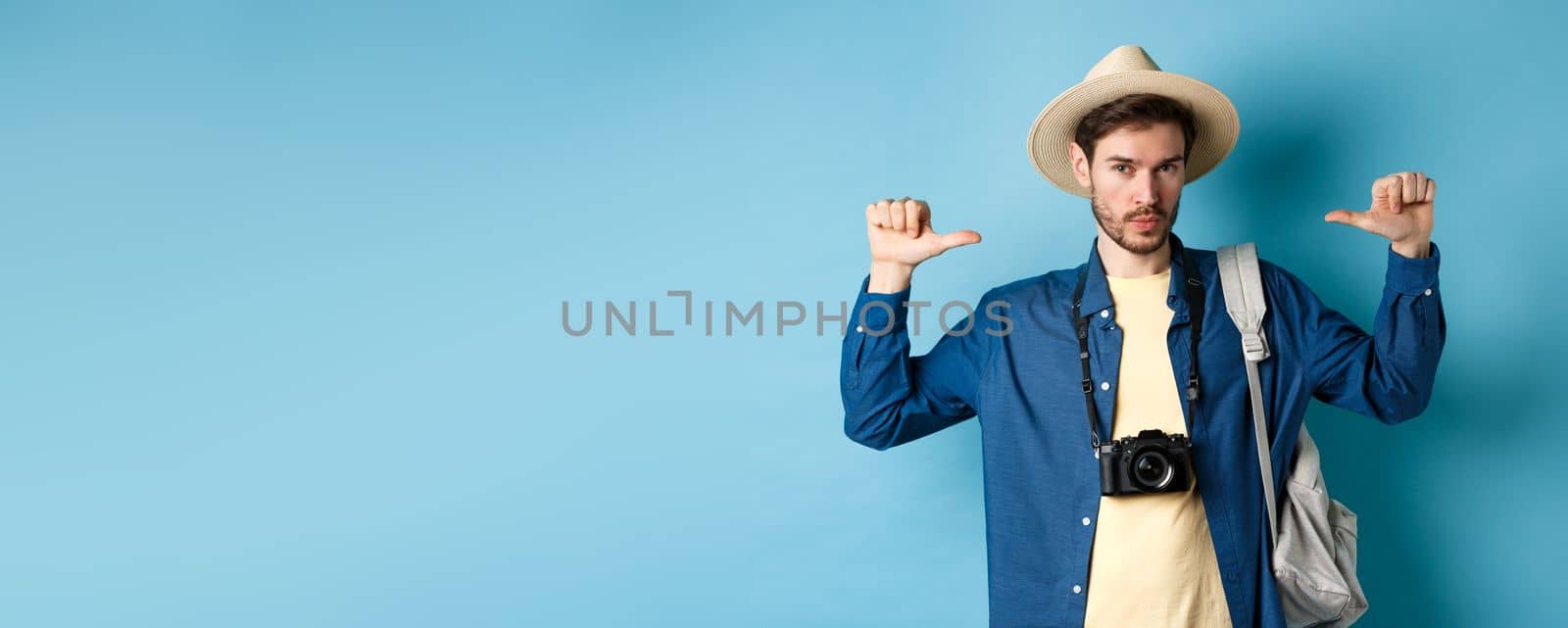 Confident handsome guy in summer hat, pointing at himself with bragging look, going to travel on holidays, holding backpack and camera, blue background.