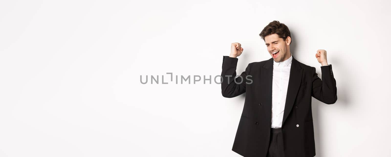 Image of successful and pleased handsome man in suit, rejoicing and making fist pump signs, dancing from happiness, standing over white background.