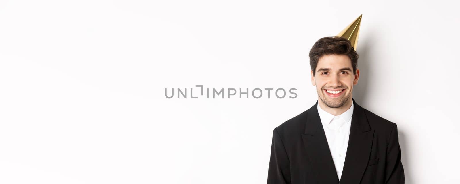 Close-up of handsome happy man in suit and party hat, smiling joyful, celebrating holiday, standing against white background.