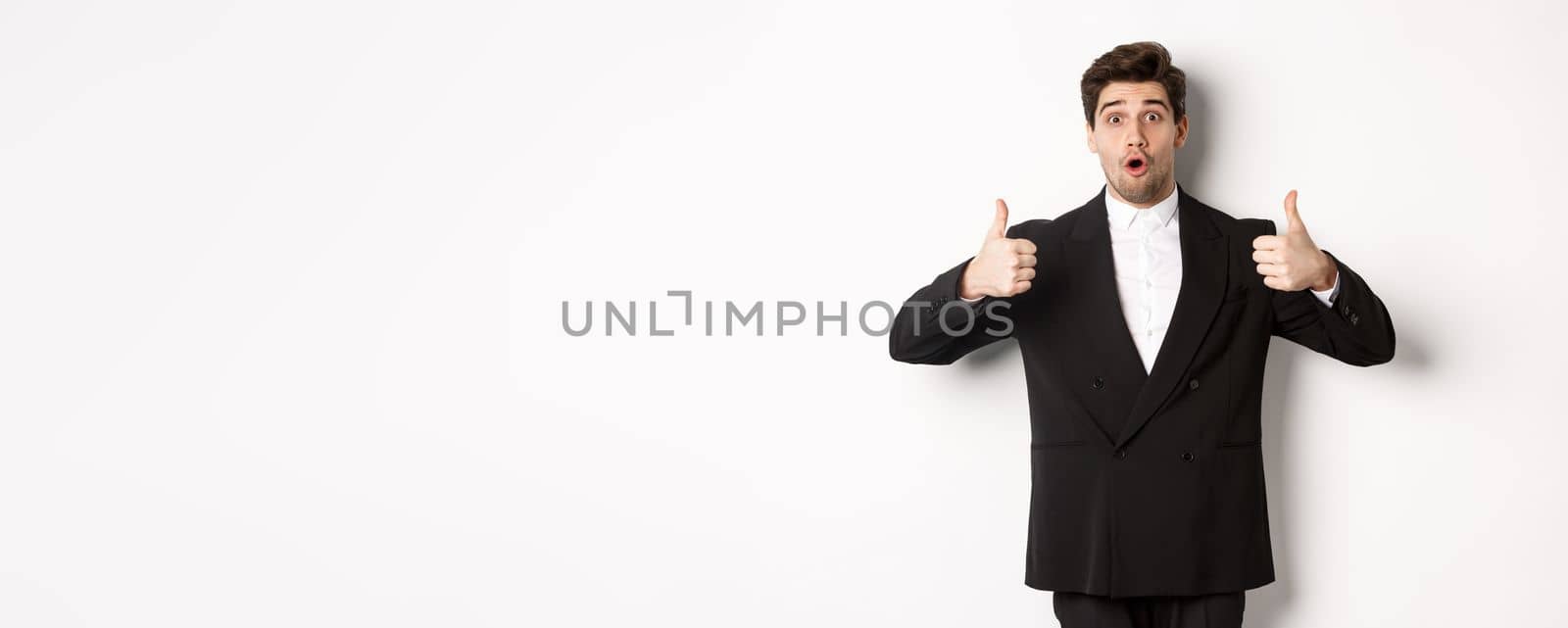 Concept of new year party, celebration and lifestyle. Portrait of excited handsome businessman in formal suit, showing thumbs-up in approval, recommending shop, standing over white background.