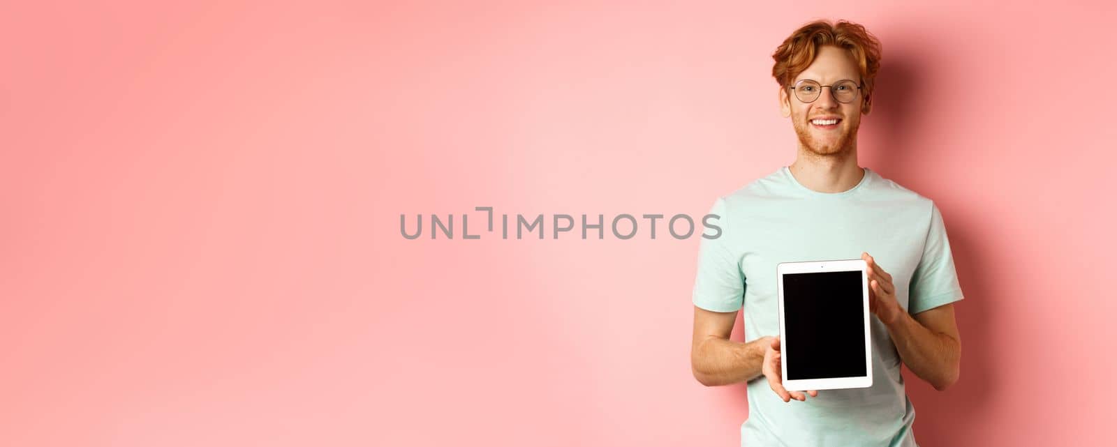 Funny redhead guy in glasses demonstrate digital tablet screen, smiling at camera, standing over pink background.