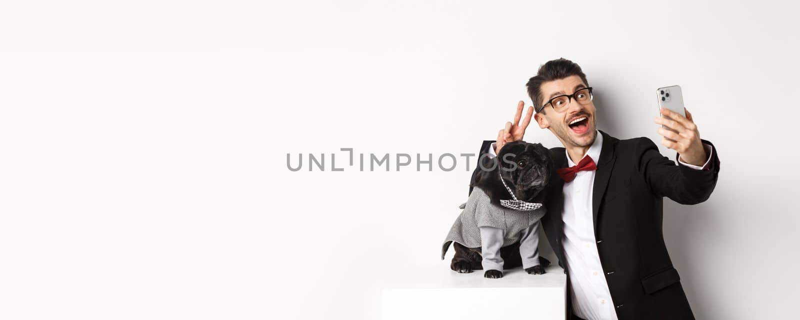 Cheerful dog owner in suit celebrating New Year with dog, taking selfie on smartphone near cute black pug in costume, white background.
