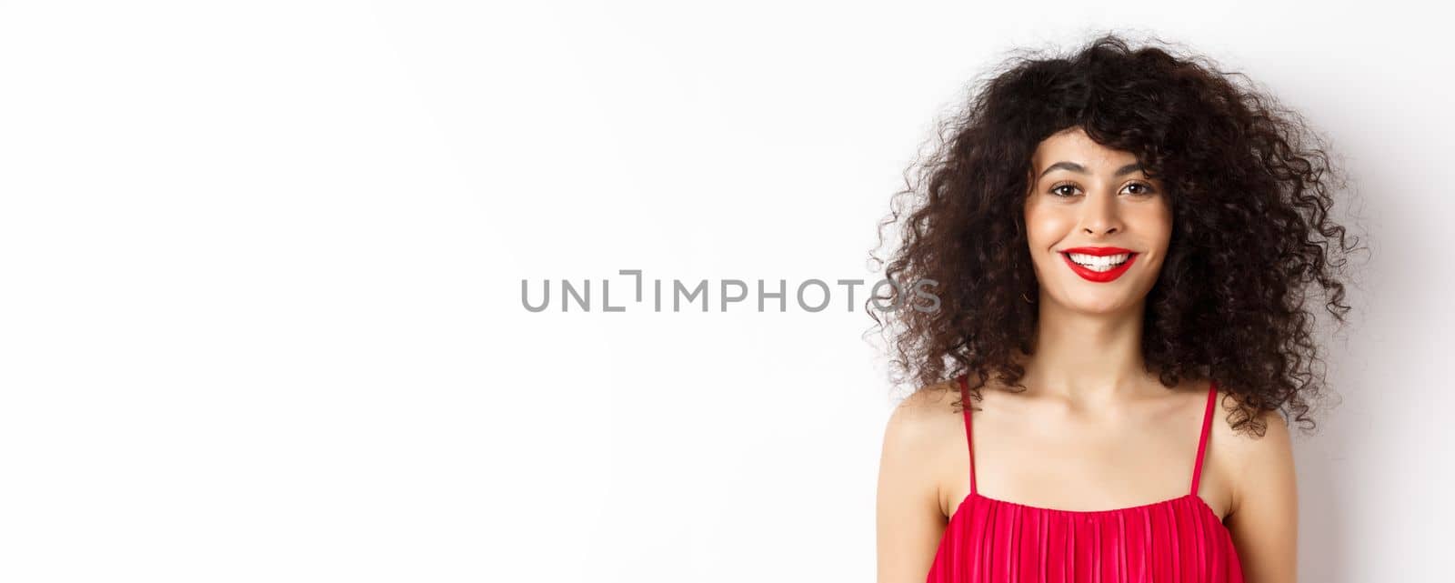 Close-up of beautiful woman with curly hairstyle, wearing red dress and lipstick, smiling happy at camera, white background.