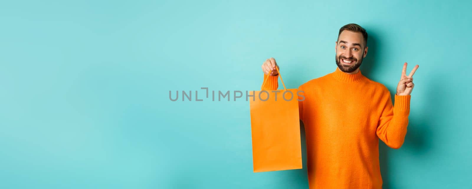 Happy young man showing peace sign and orange shopping bag, smiling pleased, standing over turquoise background. Copy space