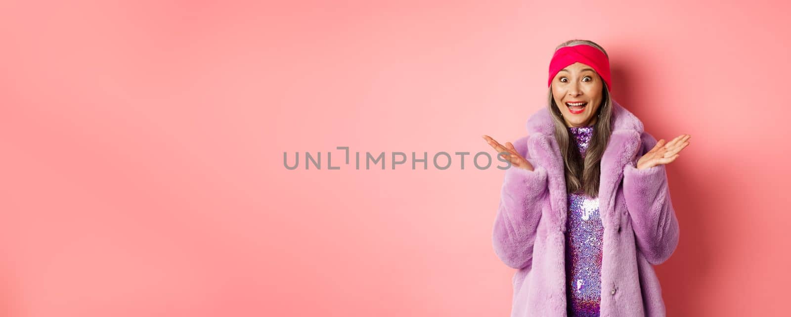 Fashion and shopping concept. Cheerful asian elderly woman in purple stylish coat looking surprised, hear good news and rejoicing, standing over pink background.