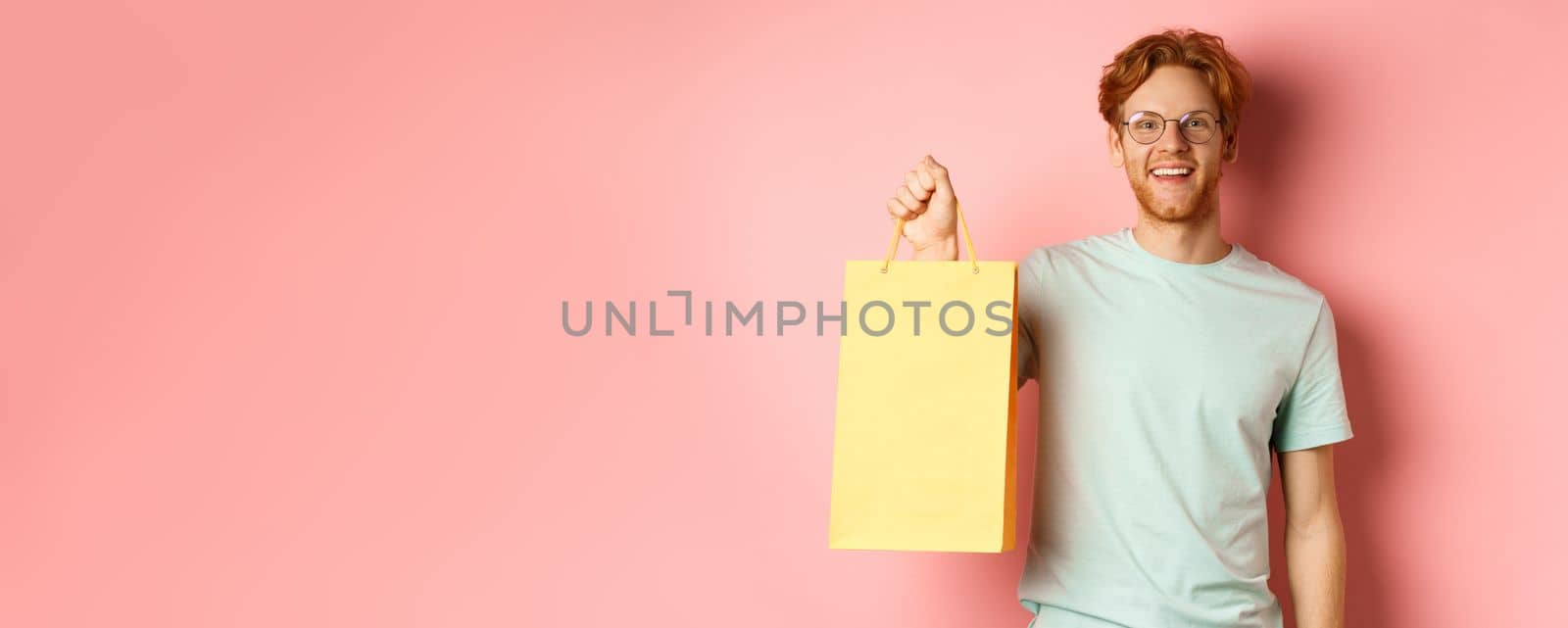 Handsome young man buying presents, holding shopping bag and smiling at camera, standing over pink background by Benzoix