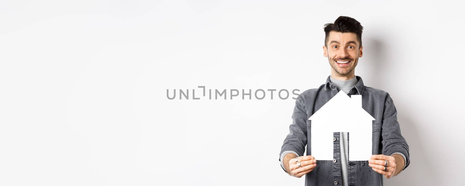 Real estate and insurance concept. Handsome young guy showing paper house cutout and smiling, searching for apartment, buying property, standing against white background.